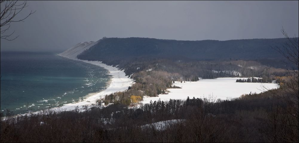 A winter snow squall approaches the Sleeping Bear Dunes while sun highlights North Bar Lake and the Empire Beach, as viewed from the lookout on the Bluffs trail.